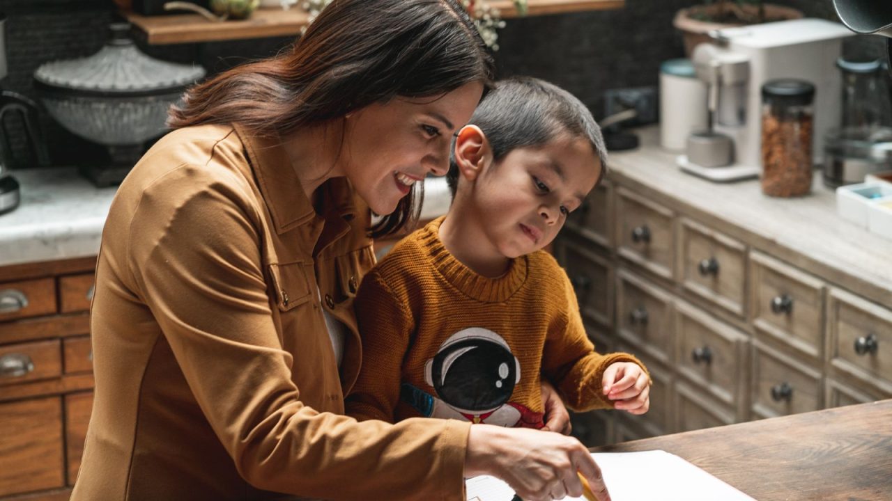 A mother in a tan shirt helps her young son in an orange sweater with homeschool work at a kitchen counter, both focused on educational materials spread on the wooden surface.