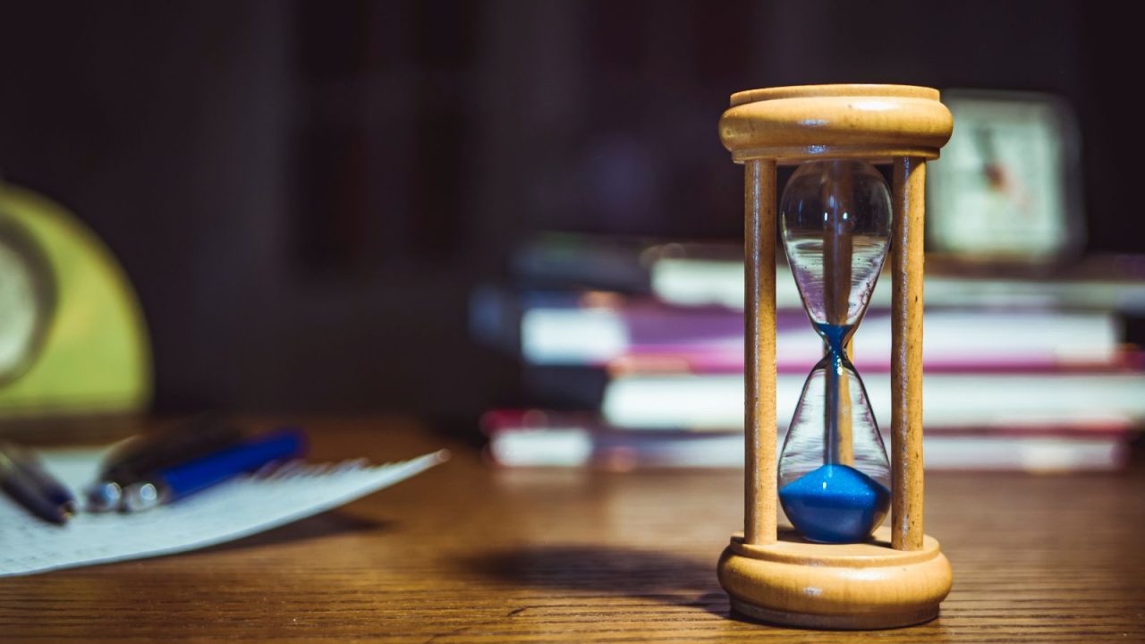 Wooden hourglass with blue sand on a desk with blurred books and papers in the background, symbolizing time management for homeschool education.