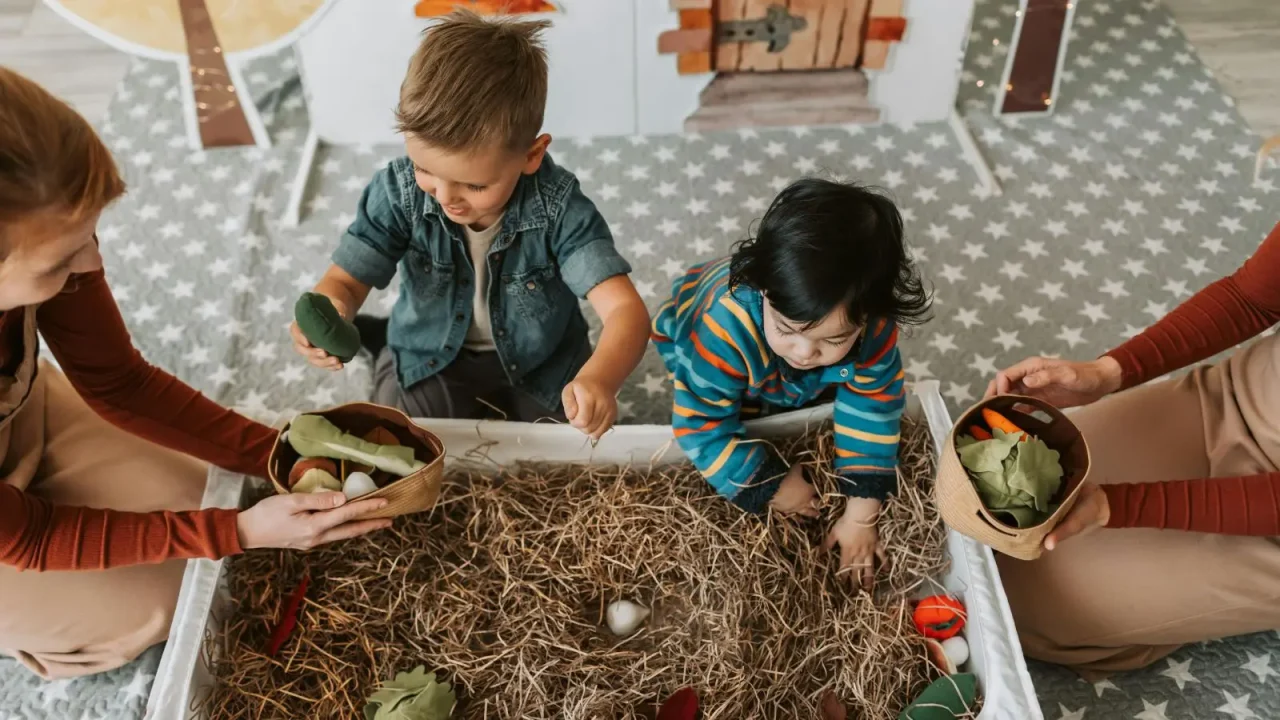 Two children exploring a natural sensory bin with straw and nest materials, guided by adults offering wooden bowls of leaves—capturing the hands-on experiential learning that makes homeschooling unique.